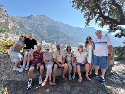 A cheerful group of travellers posing on a stone terrace with colorful hillside houses and steep coastal mountains in the background.