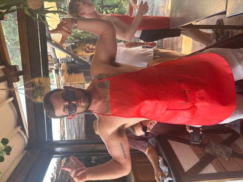 A young man wearing sunglasses and a red cooking apron flexes his arms playfully inside a rustic kitchen setting.