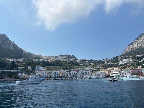 A panoramic coastal town with colorful buildings climbing the hillsides of a steep rocky island, seen from a boat on bright blue water.