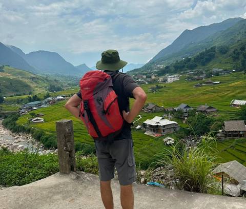A backpacker stands on a roadside railing overlooking lush rice terraces and mountain villages in northern Vietnam.