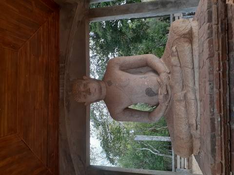 Stone seated Buddha statue sheltered under a simple pavilion with green forest in the background.
