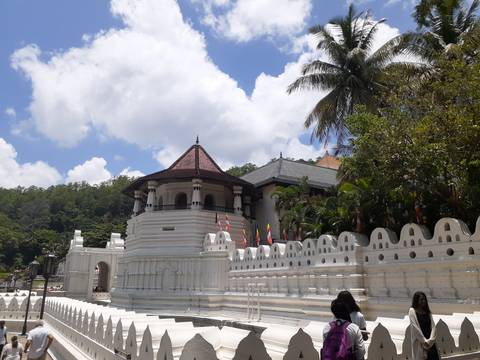 Bright midday view of the white-walled Temple of the Sacred Tooth Relic with flags and lush green hillside behind it.