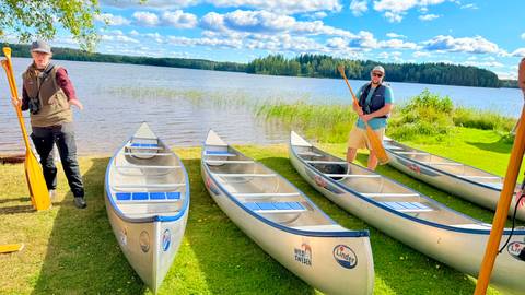 Two travelers preparing canoes on grassy lakeshore under bright blue sky.