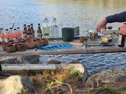 Rustic wooden table by lake set with drinks, bottles and snacks for outdoor picnic.