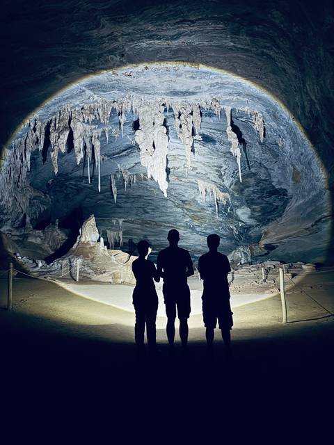 Silhouettes of three explorers facing an illuminated limestone cave interior with stalactites.