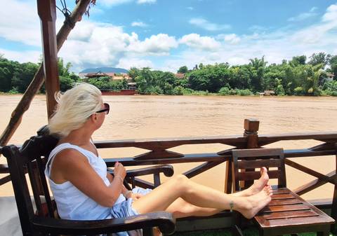 Woman relaxing on a wooden boat deck looking across a wide muddy river toward lush green banks.