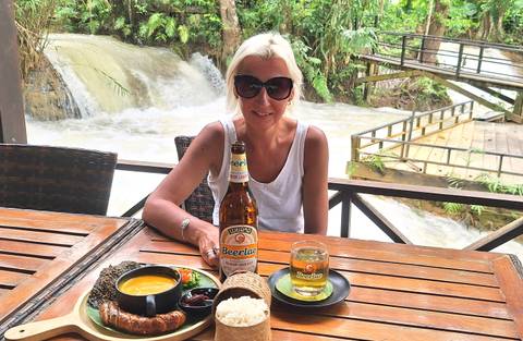 Woman seated at a wooden table enjoying Beerlao and traditional food with a waterfall backdrop.