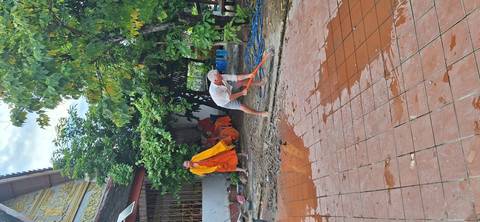 Visitor helping sweep a wet temple courtyard while orange-robed monks watch nearby.