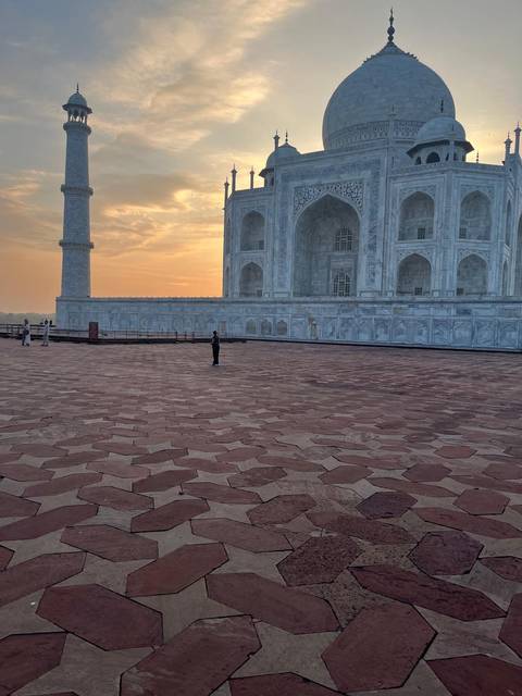 Marble platform beside the Taj Mahal at sunrise with a few visitors standing on the patterned red sandstone floor.