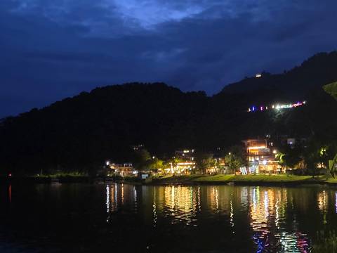 Nighttime lakeside scene with hillside lights reflecting on calm water.