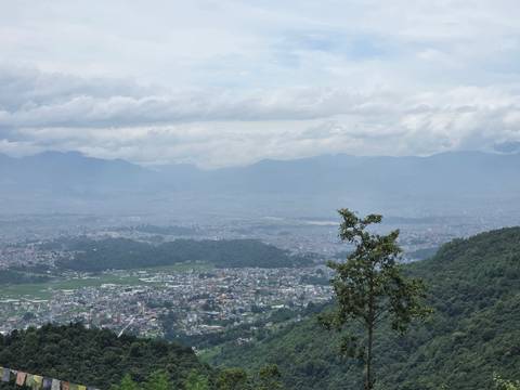 Wide view over Kathmandu Valley with cityscape, green hills and cloud-shrouded mountains beyond.