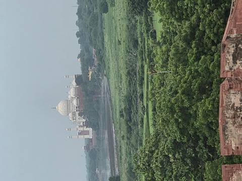 Distant view of the Taj Mahal across green plains and the Yamuna River under a hazy sky.