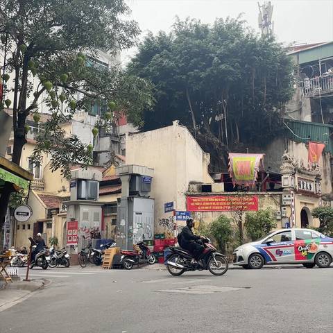 Bustling Vietnamese street corner with motorbikes, taxis, and colorful signage under leafy trees