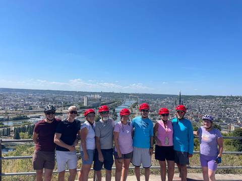 Cycling group wearing matching red helmets posing at a hilltop viewpoint over a wide river and cityscape