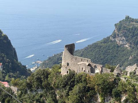 Ruined stone structure perched on a lush mountainside overlooking the Amalfi Coast and speeding boats