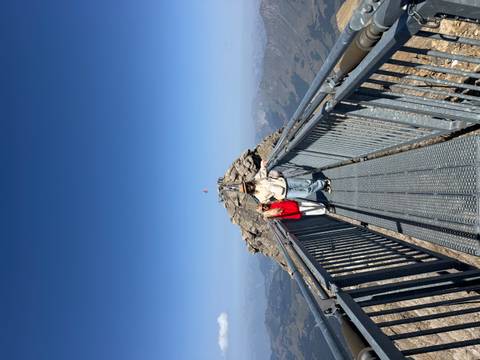 Two travelers standing on a high metal bridge walkway leading to a mountain peak with blue skies