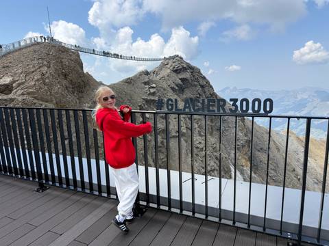 Smiling girl in red hoodie leaning on a railing with a large #GLACIER3000 sign and alpine peaks behind