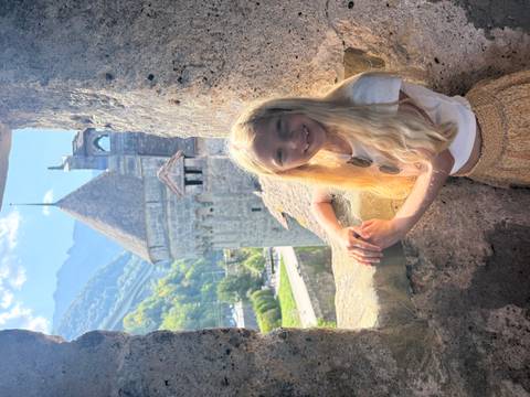 Young girl framed by stone window of a medieval castle with mountains in the background