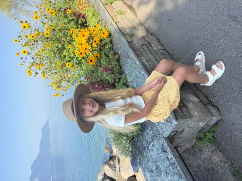 Girl with sun hat sitting beside bright yellow flowers on a lakefront promenade