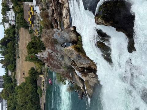 Rocky outcrop crowned with a Swiss flag at the edge of a powerful waterfall, with a couple of visitors standing on top.