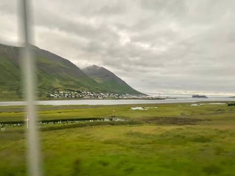 Blurry view through bus window of a coastal Icelandic town nestled below green mountains and fjord.