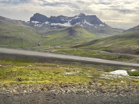 Sloping Icelandic mountain road cutting across lush valley with snow-specked peaks beyond.