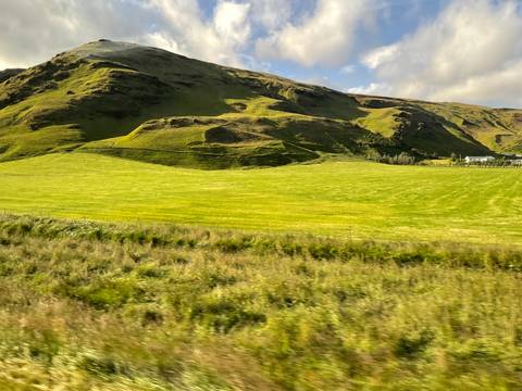 Grassy Icelandic meadow with rolling hills and farmhouse under partly sunny skies.