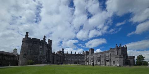 Kilkenny Castle stands majestically against a vivid blue sky filled with textured white clouds and a broad green lawn.