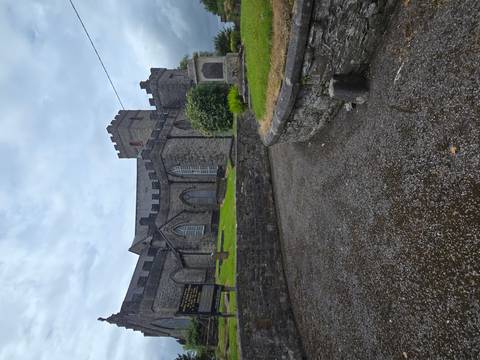 A grey-stone church with crenellated tower sits atop a damp gravel path after rainfall.