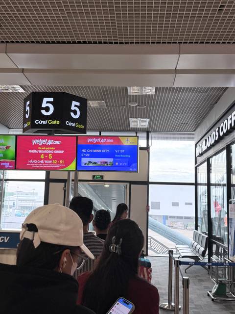Airport gate screens displaying a flight to Ho Chi Minh City with passengers waiting nearby.
