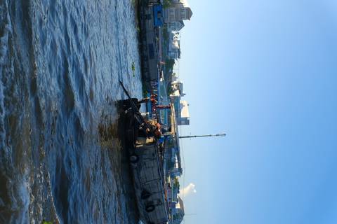 Small motorized boat navigating a bustling riverside market under clear blue skies.