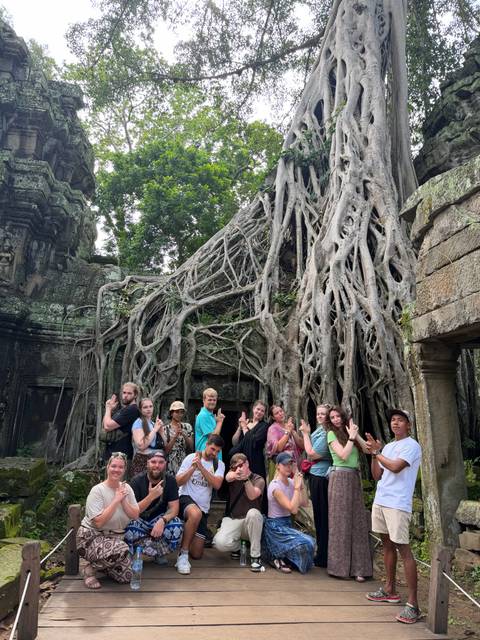 Tour group poses with playful hand gestures beneath massive tree roots enveloping ancient Angkor temple stonework.