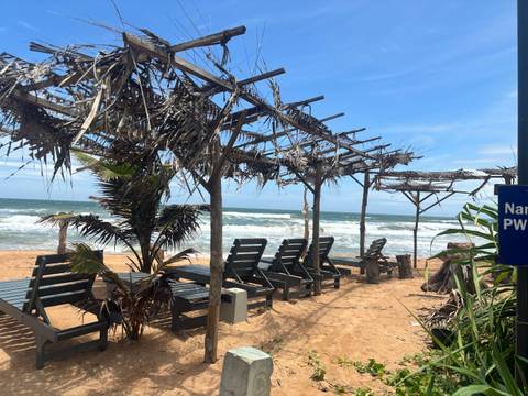 Row of empty wooden loungers beneath rustic palm-frond pergolas on a windy beachfront.