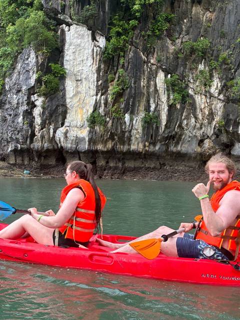 Kayakers in orange life vests paddling near limestone cliffs on emerald water giving a thumbs up.