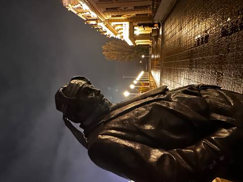 Bronze statue of a contemplative man at night on a rain-slick plaza with lit buildings behind.