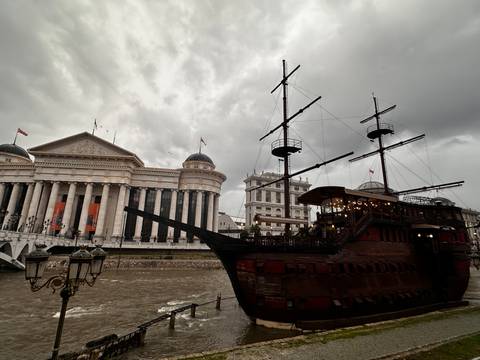 Wooden themed restaurant ship moored on river beside neoclassical museum under stormy sky.