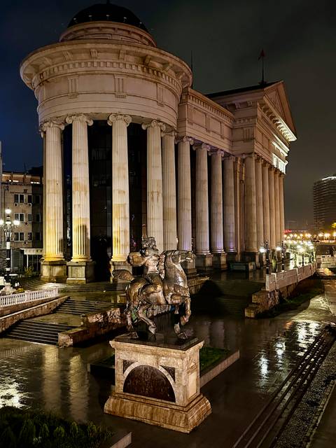 Neoclassical columned façade of the Archaeological Museum lit warmly at night with equestrian statue in front.