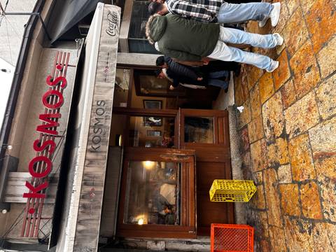 People wait outside a small eatery called Kosmos in a cobblestone alley lined with old storefronts.