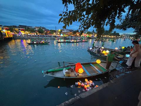 Evening river scene with lantern-lit boats and crowds along illuminated waterfront promenade.