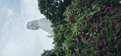 Tall white Lady Buddha statue rising above lush greenery and pink flowers against cloudy sky.