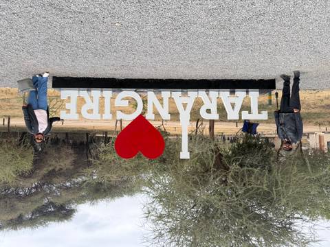 Two visitors posing beside the large 'I ♥ Tarangire' park sign in a dry woodland setting.