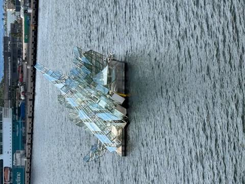 Floating glass sculpture on calm harbor water with a ferry terminal in the background.