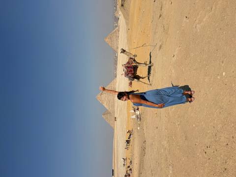 Traveller raises an arm beside a resting camel with the Giza pyramids stretching across the horizon