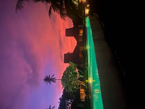 Infinity pool glowing green against vibrant pink and purple sunset skies with tropical palm and wooden villas