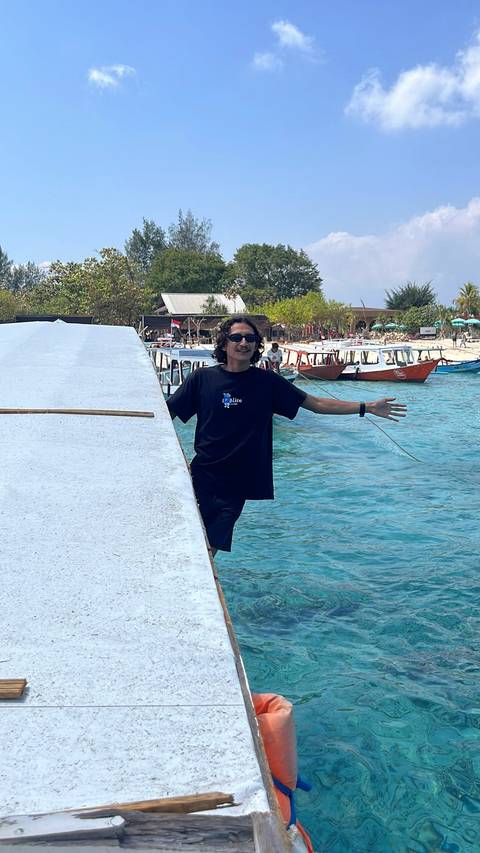 Young man spreads his arms while standing on a boat docked in bright turquoise water with island backdrop.