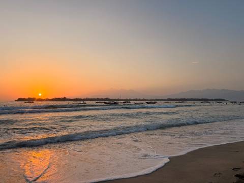 Golden sunset over gentle surf with fishing boats and distant mountains along a tranquil beach.
