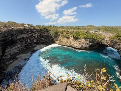 Turquoise waves crash into a horseshoe cliff bay with a natural rock arch and rugged coastline.