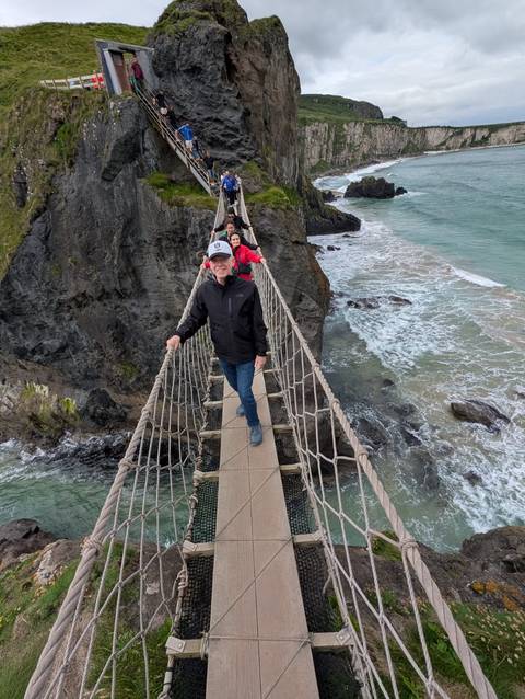 tourists carefully crossing a rope bridge high above rugged coastal cliffs and waves