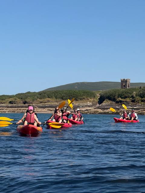 group of kayakers paddling bright red boats on calm blue water with a stone tower onshore