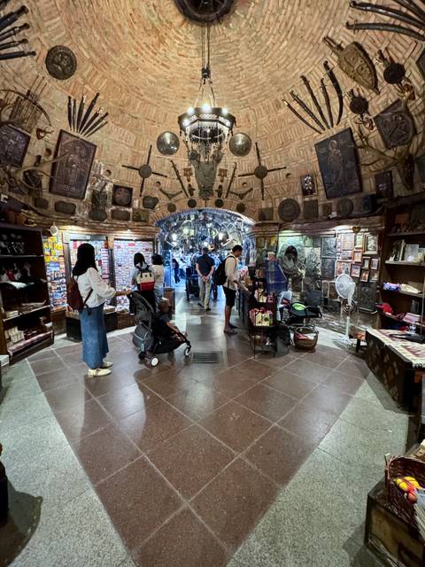 Cave-like souvenir and handicraft shop with travelers browsing shelves and carts under a decorated stone ceiling.